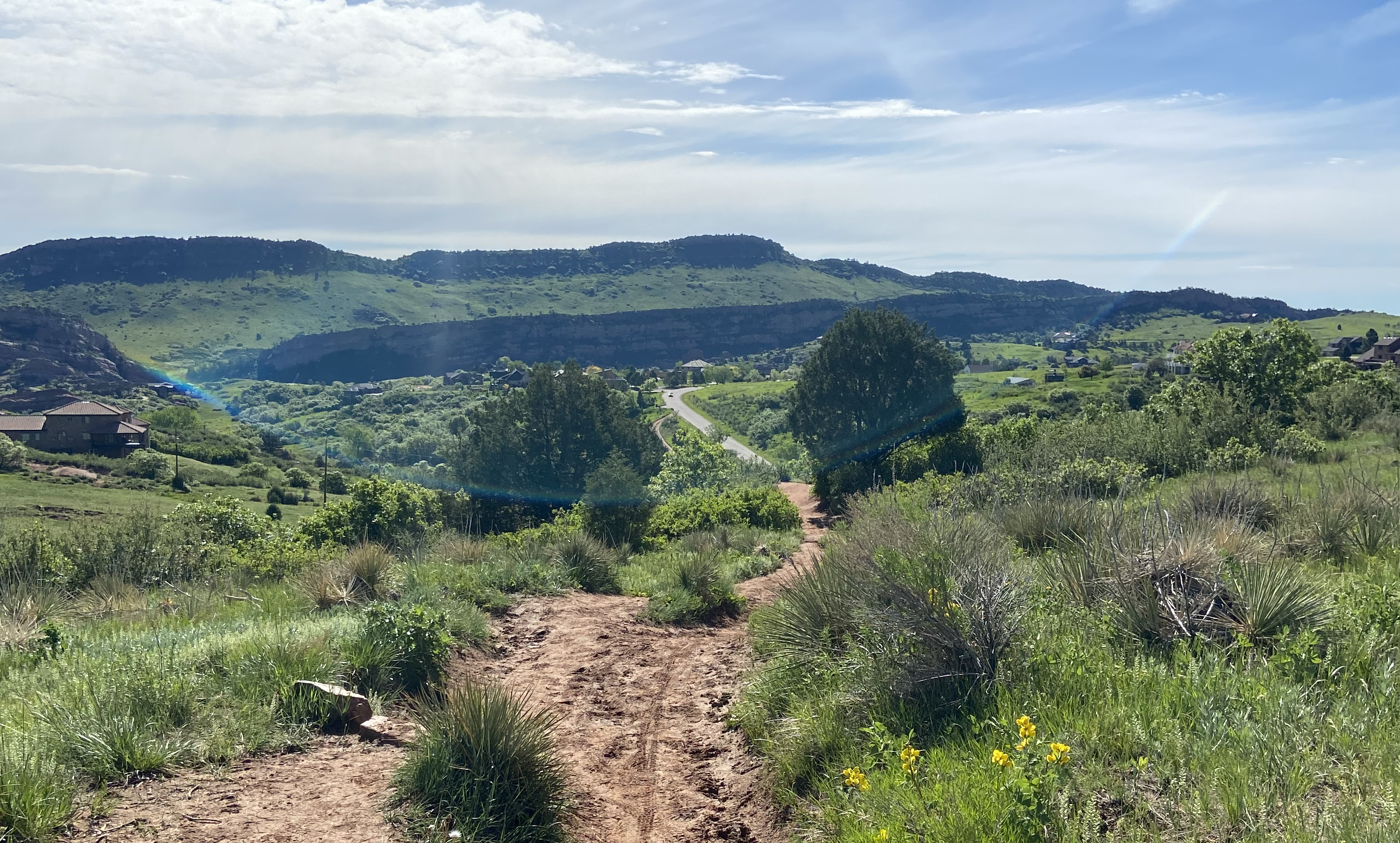 A photograph of a colorado trail surrounded in green hills and the sun shining in a blue sky.